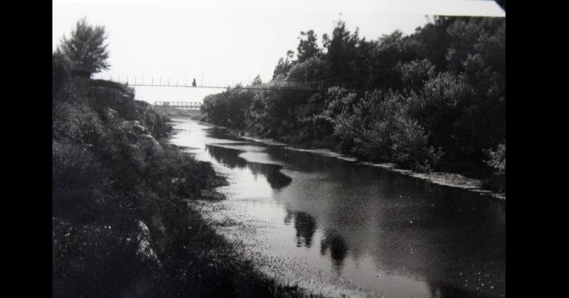 Antigua imagen donde las aguas del arroyo Tapalqué corren apacibles mientras un transeúnte cruza el puente colgante de la calle Necochea